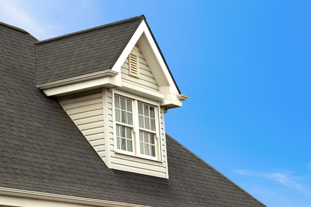Close-up of a dormer window with gray asphalt shingles against a clear blue sky.