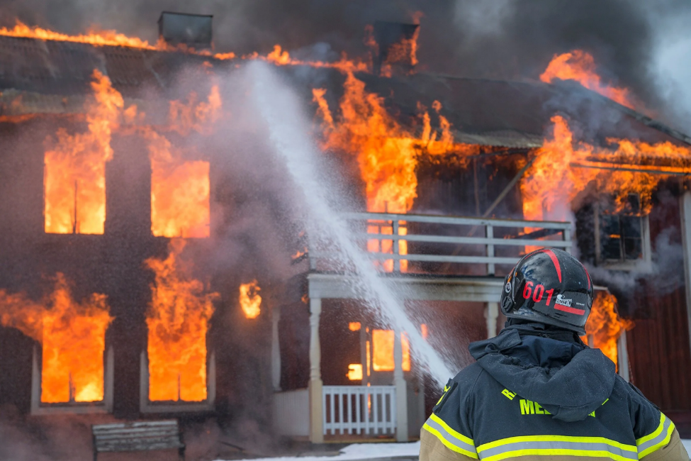 Firefighter using a hose to extinguish a house engulfed in flames.