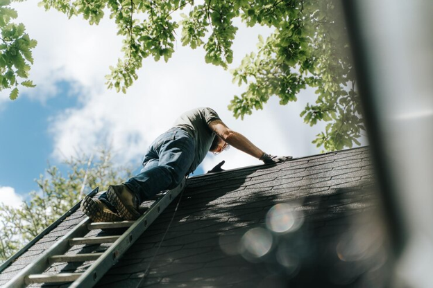 Man on a ladder installing shingles on a roof surrounded by trees.