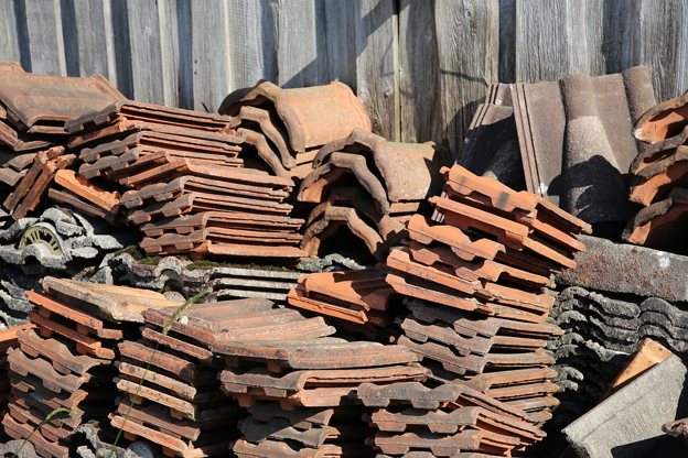 Stacks of old, broken concrete roof tiles piled against a wooden fence.